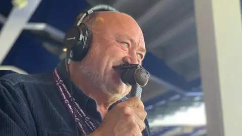 John Williams in the commentary box looking towards the pitch. He has a microphone pressed to his lips and is wearing headphones.