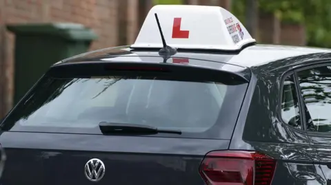 PA Media A close up photograph of a dark grey VW Polo parked on a residential street. On the roof of the car is a large white block with a big red 'L' on.