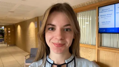 A woman with long brown hair and brown eyes is pictured looking ahead and smiling.