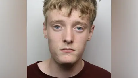 A police mugshot of a man with short blonde hair and a dark t-shirt.