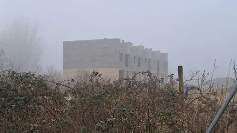 LDRS An unfinished two-storey block of flats in light and dark brick. There are no roof or windows and in the foreground are overgrown bushes 