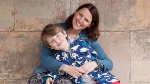 Cliff Gibson Rachel Gibson with shoulder length dark hair wearing a denim shirt with her arms around her son, aged about eight or nine, wearing a blue top with white and bright blue dinosaurs, sitting against a stone wall