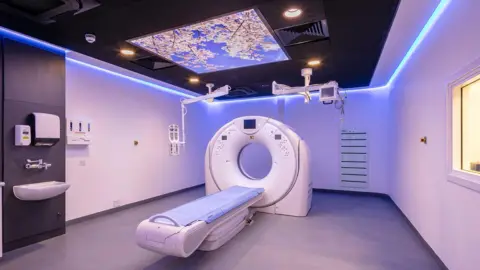 A room in a diagnostic centre. It has a flat bed and a large circular machine with a hole in it. Above the bed is a screen. On the wall is a hand basin, set of taps, a soap dispenser and a towel dispenser.