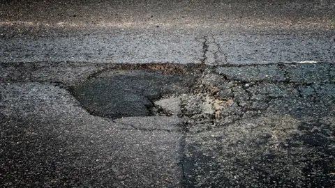 Close-up image of a pothole. It is roughly circular with a number of cracks, and part of it looks to have been filled with asphalt, which is darker in colour than the surrounding road surface.