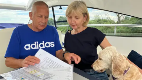 Owen Sennitt/BBC A man in a blue top and a woman in a dark blue top look at documents on a table inside a moored boat, with their dog looking on.
