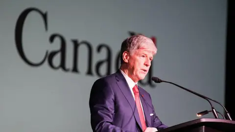 Getty Images Canada's Tim Hodgson, wearing a suit and standing in front of a microphone and lectern, with the word Canada seen on the wall behind him