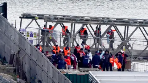 PA/GARETH FULLER A group of people who have crossed the English Channel in small boats, wearing lifejackets and being led up a gangplank to land at the the port of Dover.