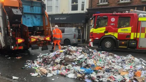 Dorset Council Waste Services The contents of a waste collection truck spilled across a street. A fire department truck is parked next to it. A worker is walking around the truck.