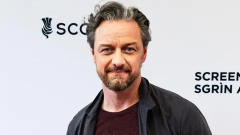 Glasgow Film Festival James McAvoy standing on the red carpet at an event. He is wearing a dark jacket, a maroon top and jeans. He had short dark hair and a beard.