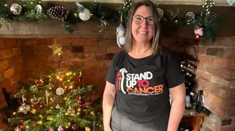 A woman with shoulder-length brown hair stands in front of a large brick fireplace with a Christmas tree in it. She is wearing glasses and a black "Stand up to Cancer" T-shirt. 