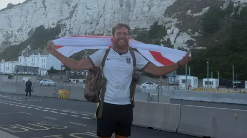 James Lewis A man with brown hair is wearing an England shift and is holding an England flag behind his back. He is standing in front of white cliffs on a road nearby.