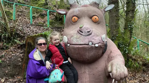 Sinead Campbell, a woman with dark hair tied back from her face, holds her four-year-old son Joshua up to a statue of the Gruffalo in a forest park. Sinead is wearing black sunglasses and a loose purple overcoat with a cream fleece-lined hood. Joshua has blonde hair tied back and is wearing a dark sweatshirt and trousers with a padded red bodywarmer. The Gruffalo is a large mythical creature with orange eyes, a wide mouth, horns, tusks and large claws.