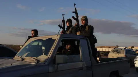 Syrian government forces hold rifles while riding on a pick-up truck on the road to Raqqa, Syria (19 January 2026)