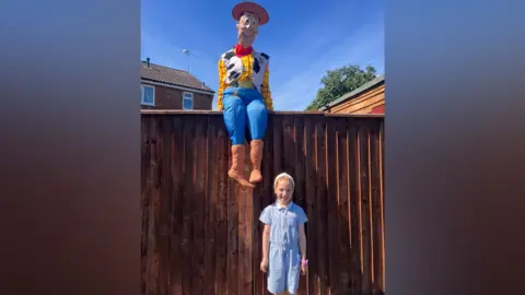Contributed A little girl wearing a blue and white school dress stands in front of a wooden fence. A life-sized sculpture of Woody from the film Toy Story is sitting on top of the fence. He is wearing a brown cowboy hat, yellow and red chequered shirt, a jacket with a cow print on it and blue trousers. 