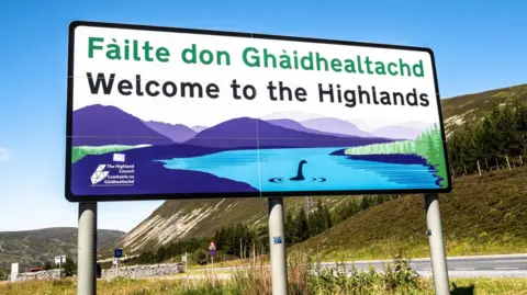 A sign on the boundary between Perth and Kinross and Highland that reads welcome to the Highlands in Gaelic and English. It features a Highland scene with Nessie in Loch Ness.