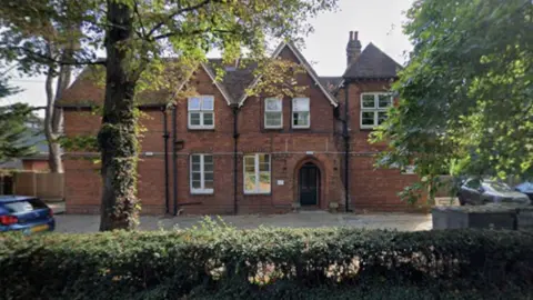 A red-brick building stands behind trees and bushes. The building has two apex roof sections and six large white-framed windows. It has a door arch for an entrance and a paved area out the front used as a car park.