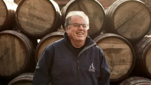 Annandale Distillery A man in glasses and a blue jacket, standing in front of a stack of whisky barrels.