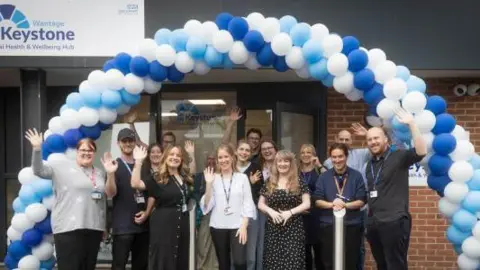 A group of people gathered together waving at the camera while stood under a blue and white balloon arch outside the front of a building