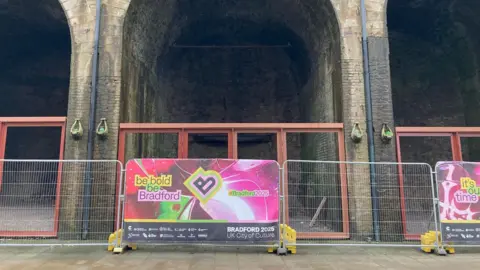 Metal fencing with a poster for Bradford City of Culture on one panel and railways arches behind with brown metal frames in them.