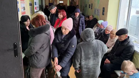 Matthew Goddard/BBC About a dozen people - of varying ages and genders - are seen in winter hats and coats as they wait in a queue at a centre for the displaced