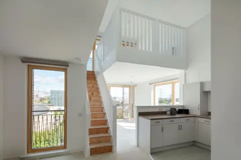 Nick Kane Apartment with a white staircase to a mezzanine level. There are windows on every wall letting loads of light in