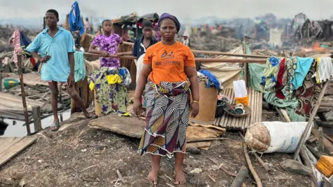 Gift Ufuoma/BBC Anna Sobie, wearing an orange T-shirt and African print fabric wrapped around her waist, stands in the ruins of her house. Her children can be seen behind her and some clothes hanging on pieces of wood.