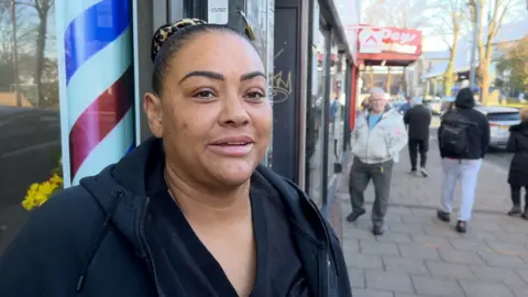 Nicola Belle, standing outside shops on a high street. She is wearing her hair in a bun and wearing a black hoodie. 