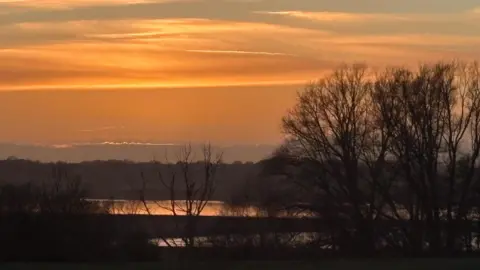 Caroline Whitworth A photo of marshland at sunset. The sky is an orange colour which is reflected in the water below. 