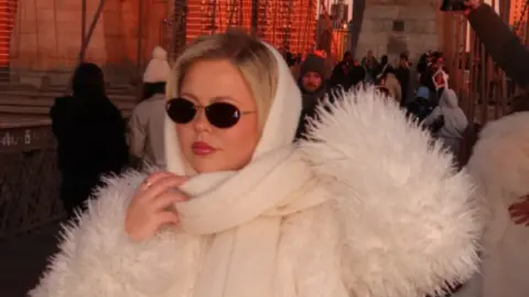 Georgia wears head-to-toe cream suit, fluffy jacket and scarf, black sunglasses. Looking chic. She has blonde hair
