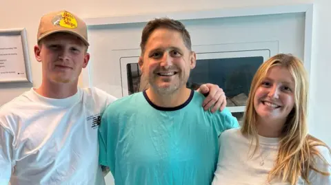 Rhys Thomas Thomas stood smiling with two of his children in hospital. He stands in the middle wearing blue hospital pyjamas. On his left, is a young man wearing a white t-shirt and beige cap. On the right, is a young woman who has long blonde hair and wears a white t-shirt. 