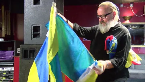 Pete with slicked back grey hair and glasses on his bearded face, unfolding a Ukrainian flag for the music event