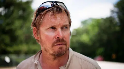 A man with sunglasses on his head and looking into the distance. He looks sun-kissed and has gingery-blonde hair with a beard.