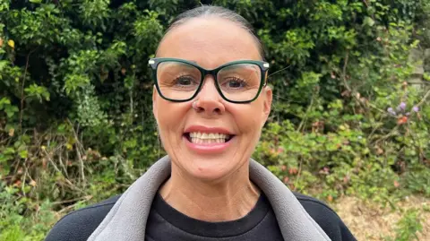 Head and shoulders image of Annette Shuttleworth. She is standing outside with greenery behind her. She is smiling, wearing large glasses and a black t-shirt and fleece. Her dark hair is tied back off her face