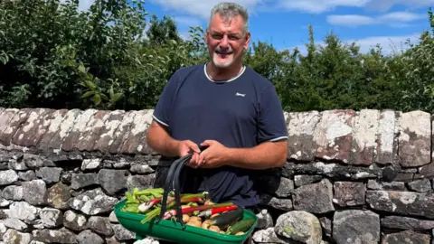 Adam Day, a man with grey hair and a beard, stands against a dry stone wall holding a basket containing vegetables from his allotment in Greystoke. He is wearing a dark t-shirt and smiling at the camera. 