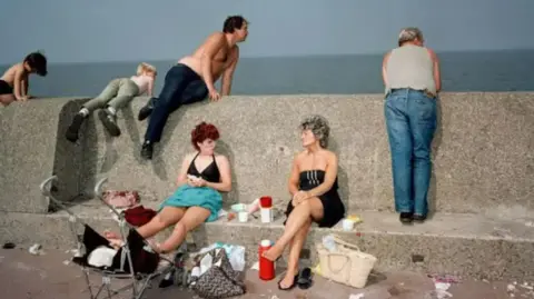 Martin Parr A photograph of New Brighton in the mid-1980s taken by photographer Martin Parr. It shows two women sitting in the foreground on the promenade wall. They are surrounded by litter. Two shirtless men lean on the wall in the background. 