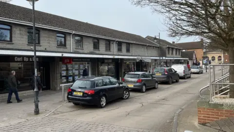 Four cars and a van parked on the side of a street in Langport. They are tucked into a parking bay outside a row of shops and an estate agent. 