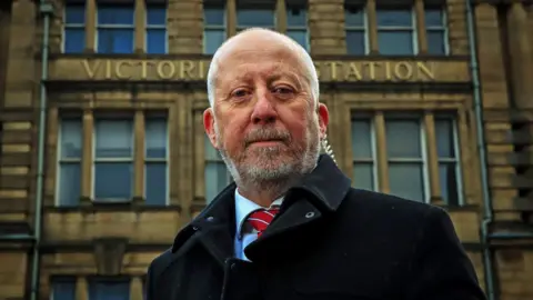 PA Media Middlesbrough and Thornaby MP Andy McDonald, wearing a black coat and red striped tie and standing outside Victoria Station, which is a large Victorian stone building with Victoria Station in gold metal letters.