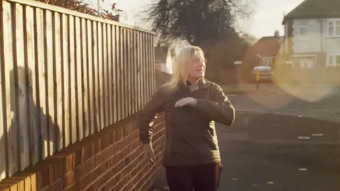 Helen Gordon in a green jacket dancing next to a fence. The sun is shining on her, reflecting her shadow against the wood.