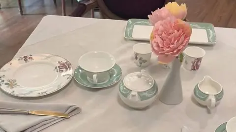 Collingtree Park Care Home A tea service in green and white including cream jug, sugar basin, tea cup and saucer. There is a red and green patterned plate and a pink flower in a vase. They are all on a white table cloth.