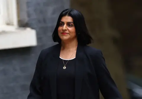 Reuters Justice Secretary Shabana Mahmood wearing a black blazer and suit, walking, and with a brick wall behind her