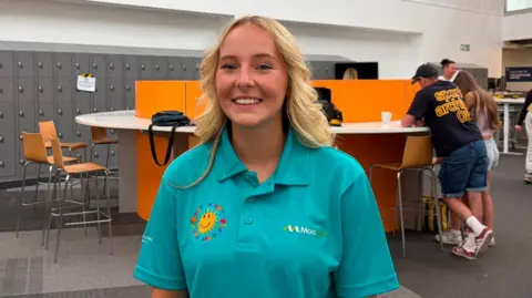 A young woman smiles for the camera as she standing in front of a desk with pupils behind her. She has shoulder length blonde hair and is wearing a teal t-shirt is a yellow sun crest 