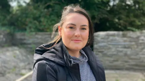 Ffion sonrió a la cámara. Ella se paró frente a una pared gris y árboles. Ffion lleva una camisa a rayas azul marino y blanca y una chaqueta negra. Ella tiene un cabello castaño oscuro que sopla en el viento.