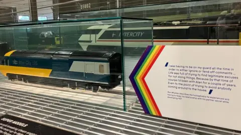 BBC/Seb Cheer A board with rainbow trim is next to models of Inter-City high speed trains. On the board, a quote reads: "I was having to be on my guard all the time in order to either ignore or fend off comments ... Life was full of trying to find legitimate excuses for not doing things. Because by that time of course I'd been with Alan for a couple of years. Even to the point of trying to avoid anybody coming around to the house." Ron Whalley describes the homophobia he experienced and having to keep his relationship with his partner secret while developing HST.