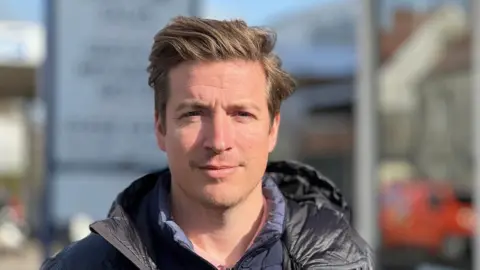 Alex Martin is wearing a coral polo shirt, a navy blue cellular jacket and a black hooded jacket