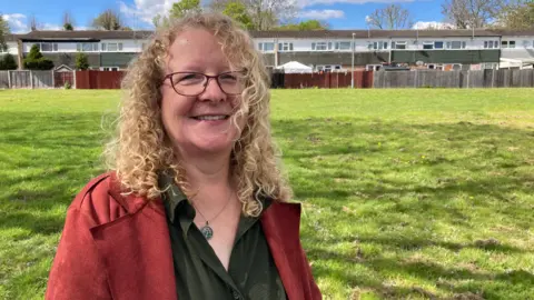 Green councillor Claire Davies is standing in a field, wearing a red jacket and green shirt.