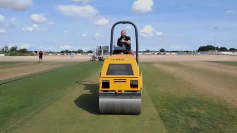 A man on a yellow cricket pitch roller with a parched outfield all around the square