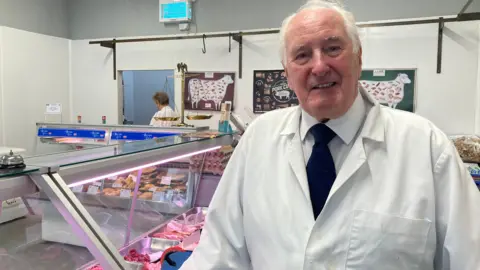 A man wearing a white apron, stood in front of a glass counter containing raw meat. 