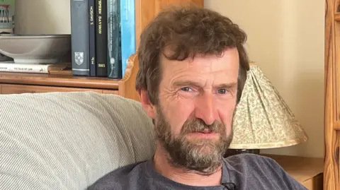 Stephen, who has a beard and short brown hair, wearing a grey top sitting on a sofa with a floral lampshade behind him in his rented home.