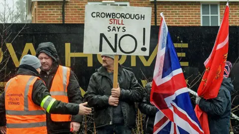 People gathered outside the Crowborough Army Camp in East Sussex waving flags and holding banners during a protest against the UK Home Office's plan. Protestesters wave union jack flags and one holds a placard which reads "Crowborough STILL says no".