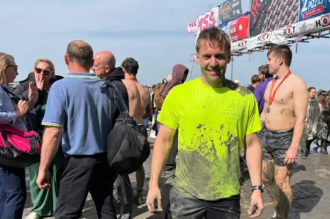 Jodie Halford/BBC A man wearing a bright yellow top which is partly covered in mud smiles at the camera. Tens of other people surround him, also covered in mud.